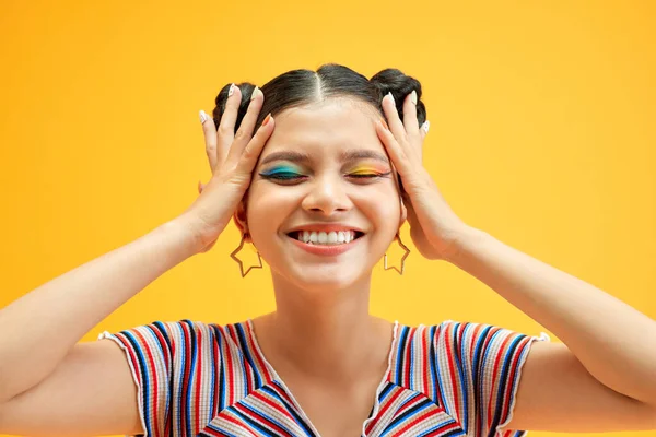 Young woman with colorful eyes smiles and covers her face with her hands, posing on yellow background