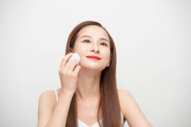 Beyaz arka planda izole edilmiş, makyaj süngeri kullanan güzel bir genç kadının portresi.