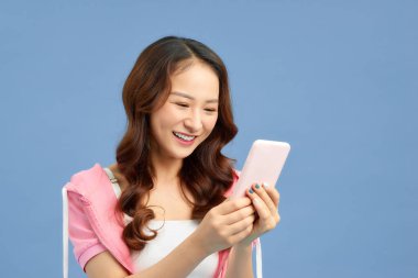 Akıllı telefonuyla mesaj atan mutlu genç Asyalı kadın mavi arka planda..