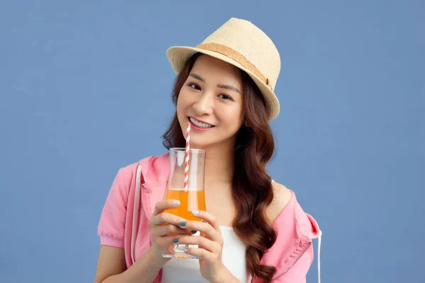 幸せな若いアジアの女性の肖像画を身に着けている藁帽子と青の背景にフルーツジュースを飲みます — ストック写真