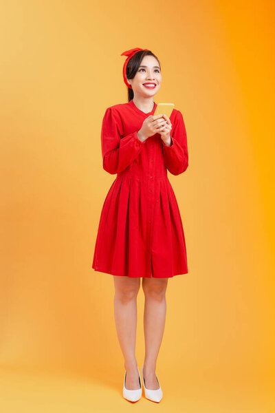 Happy young Asian woman in red dress using smartphone and standing isolated over orange background.