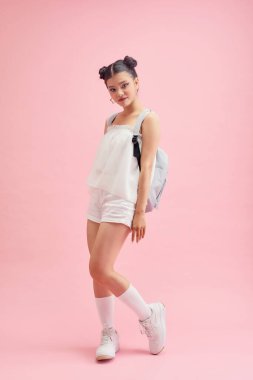 Moda okullu kız sırtında bir sırt çantası tutuyor ve pembe arka planda izole edilmiş kameraya bakıyor. 