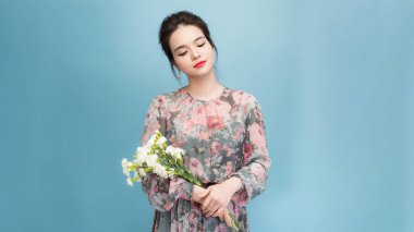 Hoş görünümlü kadın portresi az makyajlı, geniş gülümsemeli, özel durumlarda çiçek alan, açık mavi arka planda izole edilmiş.