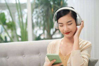 Gülümseyen kız evde dinleniyor, akıllı telefon kullanıyor ve beyaz kulaklık takıyor.