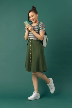 Asyalı neşeli üniversite öğrencisi yeşil arka planda izole akıllı telefon kullanıyor.