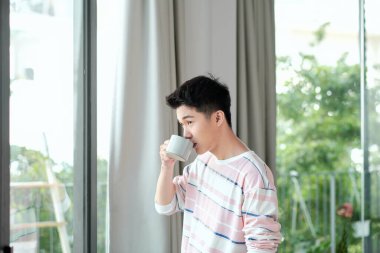 Genç yakışıklı adam evinin penceresinde sabah kahvesini içiyor.