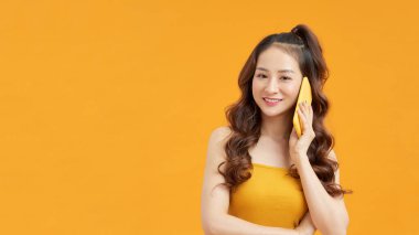 Güzel gülümseyen Asyalı bir kadının görüntüsü. Sarı duvar arkasında izole edilmiş poz veriyor. Cep telefonuyla konuşuyor..