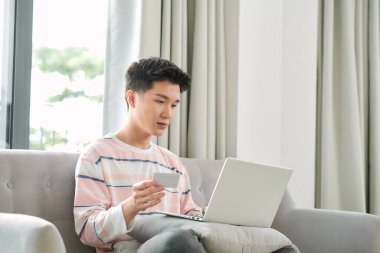 Evdeki koltukta dizüstü bilgisayarla oturan ve internetten alışveriş yapmak için kredi kartı kullanan genç etnik kökenli adamın profili.