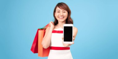 Renkli kâğıt torbalı mutlu bayan, boş ekranlı akıllı telefonu gösteriyor ve mavi arkaplanda izole edilmiş siparişi alıyor.