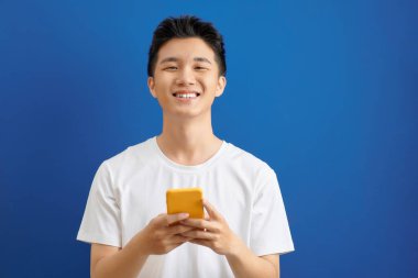 Sıradan giyinmiş öğrenci mavi arka planda izole bir şekilde duruyor akıllı telefon ekranına bakıyor, web sayfalarına bakıyor ve arkadaşlarıyla sohbet ederken gülümsüyor.