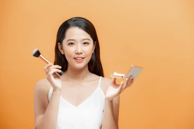 Güzel Asyalı genç kadın gülümsüyor ve makyaj fırçasıyla sağlıklı, temiz ve taze teniyle çok mutlu ve neşeli hissediyor.,