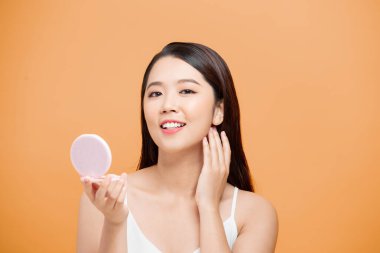 Güzel Asyalı kadın, sarı arka plandaki aynada tenine bakıyor.