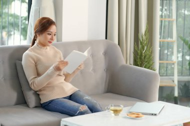Oturma odasında elinde çayla kitap okuyan bir kadın.
