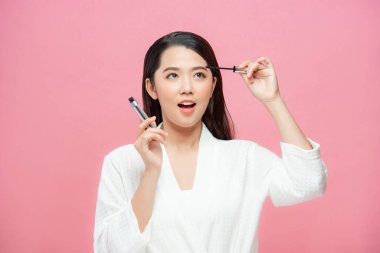 Asyalı bir kadın pembe arka planda kirpiklere rimel sürüyor.