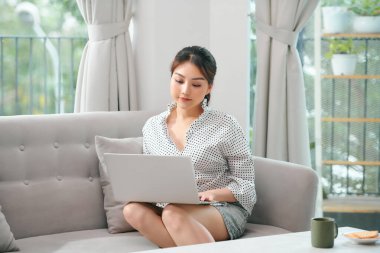 Oturma odasında kanepede otururken dizüstü bilgisayar kullanan neşeli Asyalı kadın görüntüsü