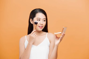 Güzel Asyalı genç kadın gülümsüyor ve makyaj fırçasıyla sağlıklı, temiz ve taze teniyle çok mutlu ve neşeli hissediyor.,
