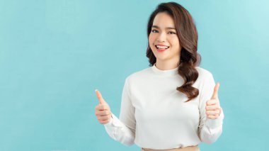 Gülümseyen güzel bir genç kadın mavi arka planda baş parmak gösteriyor.
