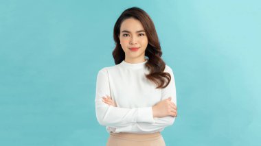 Mavi arka planda kolları çapraz dururken kameraya gülümseyen basit tişörtlü neşeli bir kadın görüntüsü