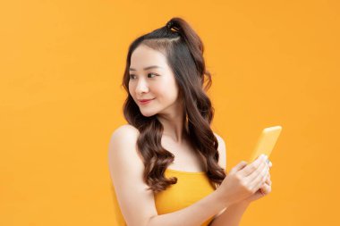 Asyalı bir kız telefonu tutar ve sarı arka plana bakar..