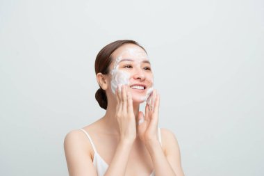 Tatmin olmuş güzel bir kadın yüz masajı yapıyor, beyaz köpüğe sahip, cilt bakımlı, gülümseyen, beyaz stüdyo duvarının üstünde mankenler.