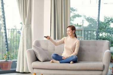 Olumlu gülümseyen iyimser bir genç kadının evde oturup elinde uzaktan kumandayla televizyon izlemesi..