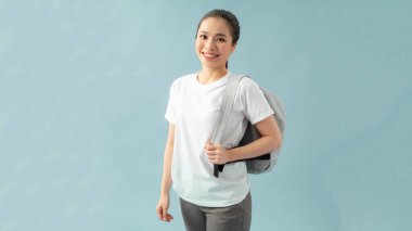 İzole edilmiş mavi arka planda duran sırt çantası takan öğrenci kadın kameraya bakıp gülümsüyor..