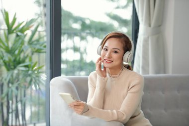 Gülümseyen kız evde dinleniyor, akıllı telefon kullanıyor ve beyaz kulaklık takıyor.