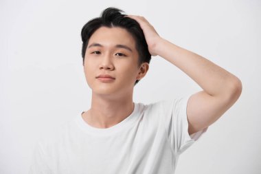 İzole edilmiş arka planda beyaz tişört giyen genç yakışıklı adam. Saçına dokunup el hareketleriyle gülümsüyor.