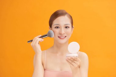 Güzel genç bir kadın makyaj fırçası ve küçük ayna kullanarak yanağına allık sürüyor.