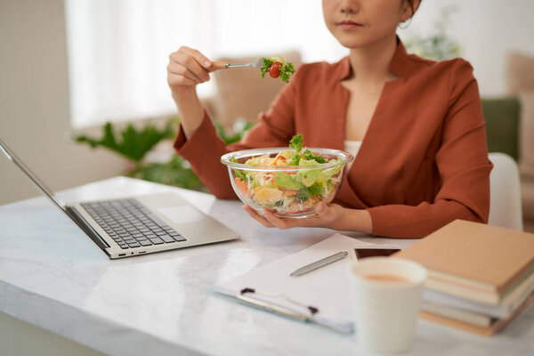 woman with laptop in modern office interior, eating lunch.