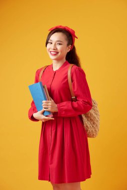 Sırt çantalı güzel bir kız öğrenci, sarı bir arka planda izole edilmiş defterleri elinde tutuyor.