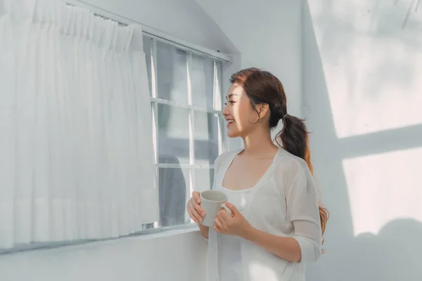 Joven Mujer Sonriente Mirando Por Ventana Con Una Taza Bebida — Foto de Stock