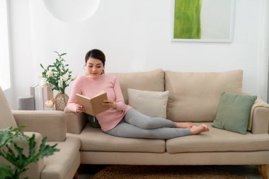 Güzel, gülümseyen bir kadın kitap okuyor ve oturma odasındaki kanepede uzanıyor.