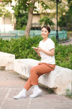 Şehir parkında bir bankta oturan akıllı telefonu kullanan mutlu kız.