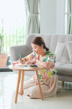 Happy Wonam evde yerde oturmuş deftere notlar yazıyor.