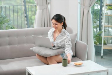 Oturma odasında kanepede otururken dizüstü bilgisayar kullanan neşeli Asyalı kadın görüntüsü
