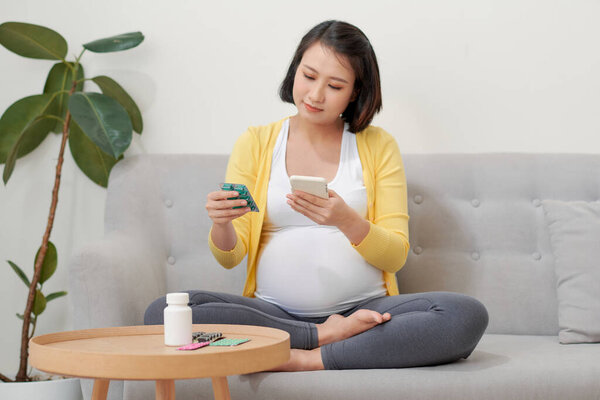 Asian pregnant woman using mobile phone searching information and reading medicine label and prescription medications