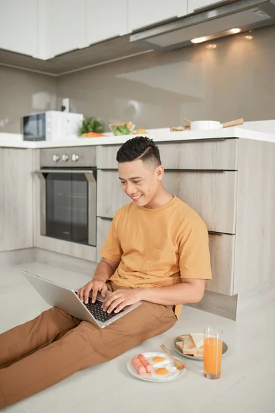 英俊的男性坐在地板上 膝头抱着笔记本电脑 一边吃早饭 — 图库照片