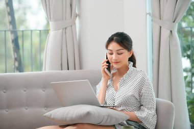 Evde dizüstü bilgisayarlı bir koltukta otururken cep telefonu kullanan mutlu genç Asyalı kadın.