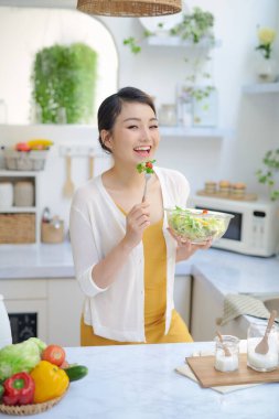 Mutfakta sebze salatası yiyen gülümseyen genç bir kadın.