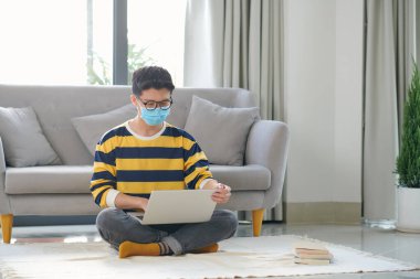 Asyalı adam elinde bir kitap ve dizüstü bilgisayarla yerde oturuyor.