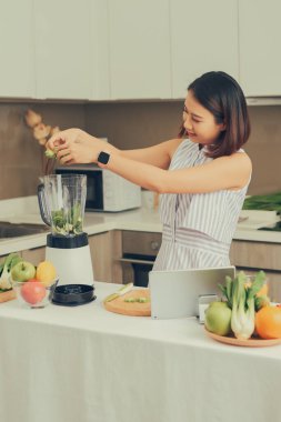 Gülümseyen vejetaryen Asyalı kadın mutfakta meyve ve sebzeyle smoothie yapıyor.
