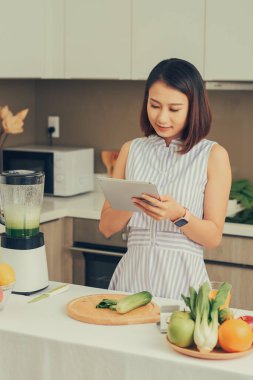 Asyalı bir kadın mutfakta meyve ve sebzeli smoothie yapmak için tablet kullanıyor..