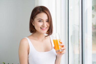 Genç neşeli kadın portakal suyu içiyor ve pencerenin yanında duruyor.