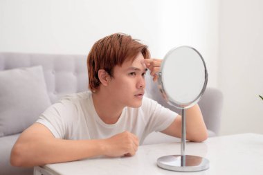 Genç yakışıklı Asyalı adam aynada yalnız başına yüzüne bakıyor.