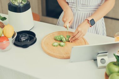 Asyalı kadın önünde tabletle mutfakta sebze kesiyor.