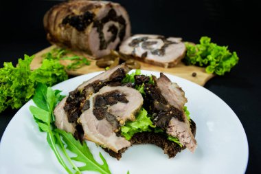 Etli, otlu, salata yapraklı ve roka aromalı sandviç. Siyah arka planda beyaz bir tabakta atıştırmak için. Kuru erik ve mantarlı domuz eti dilimlenmiş tahtada..