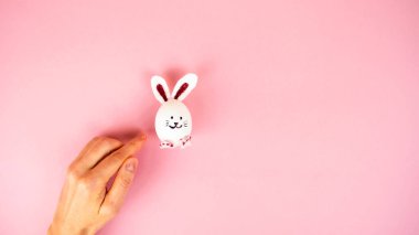 Female hand near a cute Easter bunny made from a white chicken egg isolated on a light pink pastel background. Happy Easter Day. Greeting card. Copy space. Template for design and text.