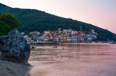 Pembe günbatımı ışınlarıyla tepedeki eski köyün güzel manzarası. Moscenicka Draga, Hırvatistan. Çakıl taşı Sipar plajından görüntü. Adriyatik Denizi.