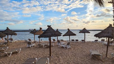 Gün batımında palmiye ağaçları, güneş ışıkları ve şemsiyeleri olan güzel, geniş kumlu bir sahil. Playa de las Americas, Tenerife, İspanya. Atlantik Okyanusu.
